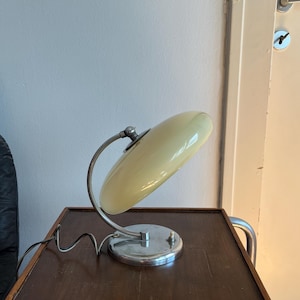 May include: A table lamp featuring a pale yellow, disc-shaped glass shade supported by a curved, silver-toned metal arm. The lamp has a circular chrome base with a small white push-button switch and a black power cord extending from the side.