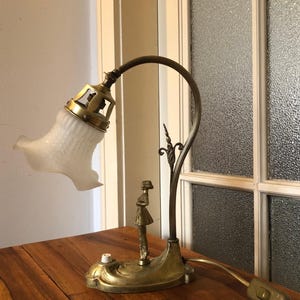 May include: A vintage brass desk lamp with a white frosted glass shade and a decorative figure of a woman. The lamp has a flexible arm and a switch on the cord.