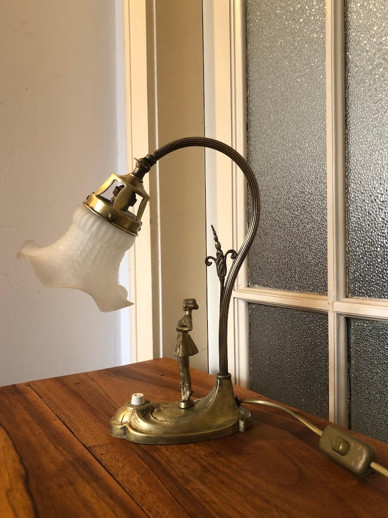 May include: A vintage brass desk lamp with a white frosted glass shade and a decorative figure of a woman. The lamp has a flexible arm and a switch on the cord.