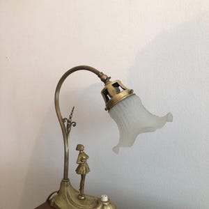 May include: A brass desk lamp with a white frosted glass shade. The lamp features a decorative figure of a woman standing on the base.