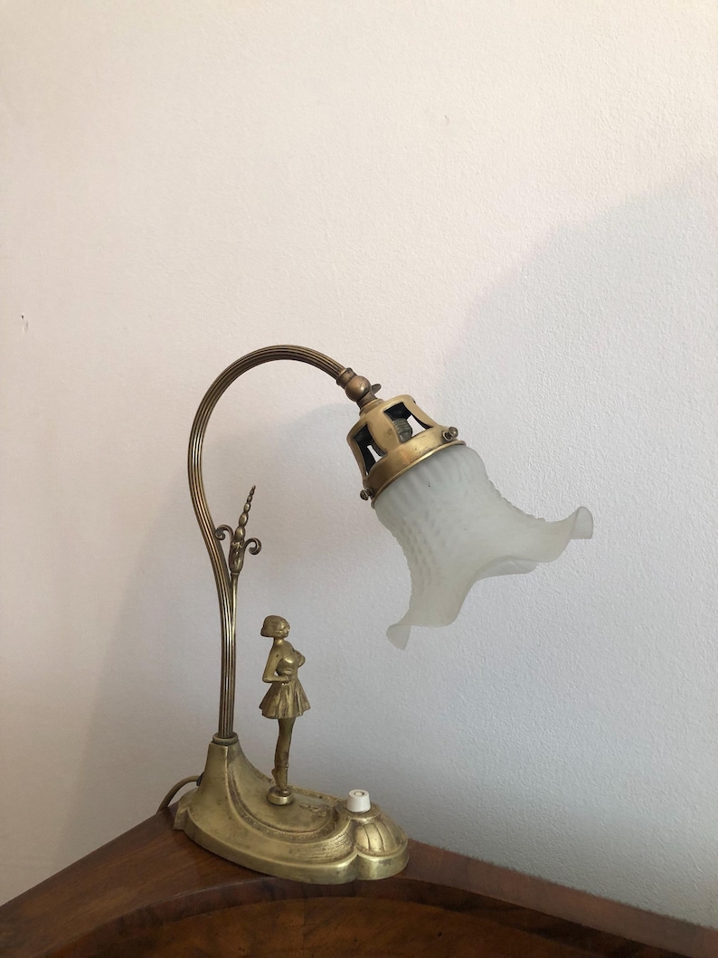 May include: A brass desk lamp with a white frosted glass shade. The lamp features a decorative figure of a woman standing on the base.