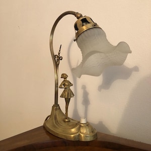 May include: A brass table lamp with a frosted glass shade shaped like a flower. The lamp features a sculpted figure of a woman standing on a base.