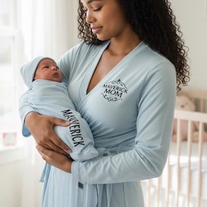 Peut inclure: Ensemble de robe et de couverture pour bébé bleu clair. La robe porte l'inscription "MAVERICK MOM" et la couverture pour bébé porte le nom "MAVERICK". Le bébé porte un bonnet bleu clair assorti.