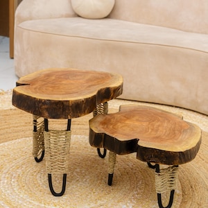 May include: Two wooden side tables with live edges. The tables have a natural wood grain finish and are supported by black metal legs wrapped with woven rope. The tables are placed on a round jute rug.