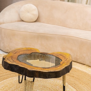May include: A unique, handcrafted coffee table with a live edge wood top and a glass insert. The table has three black metal legs wrapped with woven rope. The table is set on a round jute rug, with a beige sofa in the background.