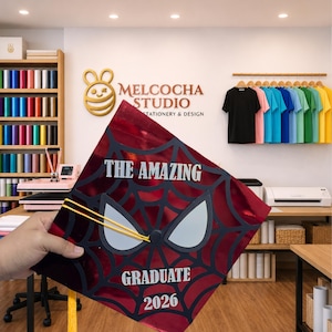 May include: A red and black graduation cap with a Spider-Man design, the words "THE AMAZING GRADUATE 2026" are printed on the cap. The cap has a yellow tassel. The image also shows a stationery and design studio.