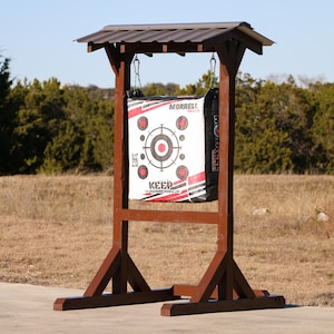 May include: A brown wooden archery target stand with a red and white archery target hanging from it. The target has the words "Morrell" and "Keep Hammering" printed on it.