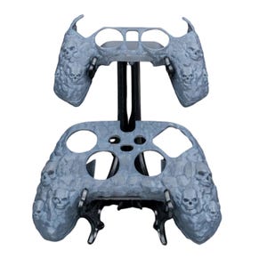 May include: A gray and black skull-patterned controller skin for a gaming console. The skin is made of a textured material and features a skull design.