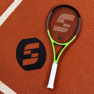 Op de afbeelding: Een groene en zwarte tennisracket met een witte grip ligt op een roodbruine baan. Een zwart logo staat naast het racket. Het racket heeft een wit snarenpatroon en de merknaam "Wilson".