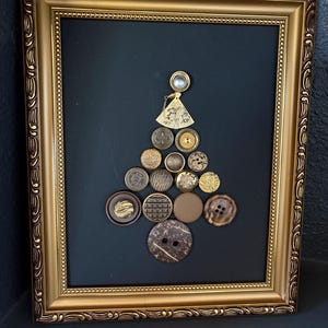 May include: A framed artwork depicting a Christmas tree made from assorted buttons. The buttons are in gold, brown, and beige tones, set against a dark background. The frame has an ornate gold finish.