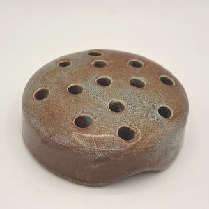 May include: A brown ceramic flower frog with multiple holes for arranging stems. The frog has a rounded shape with a speckled, glazed finish in shades of brown and blue. The holes are evenly spaced across the top surface.