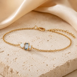 May include: A gold-tone chain bracelet featuring a central cluster of three stones: a round white opal, a rectangular light blue gemstone, and a small clear round stone. The bracelet includes a delicate link chain and a lobster claw clasp.