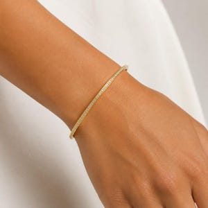 Puede incluir: Una pulsera rígida fina de tono dorado que presenta una hilera continua de pequeñas piedras claras y brillantes engastadas a lo largo de la superficie exterior. La pulsera tiene un acabado liso y pulido con un diseño circular y delgado.