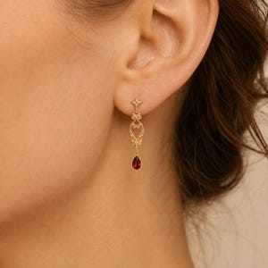 Peut inclure: Une boucle d'oreille pendante en or avec un motif en filigrane métallique orné et symétrique sur le dessus, relié à une petite pierre précieuse rouge foncé en forme de goutte qui pend. La boucle est fixée au lobe par un fermoir à tige.