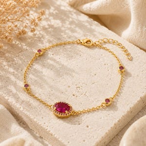 May include: A gold-tone chain bracelet featuring a central oval-shaped pink gemstone surrounded by a halo of tiny pink stones. The bracelet includes four additional small, round pink stones set along the chain and a lobster claw clasp closure.