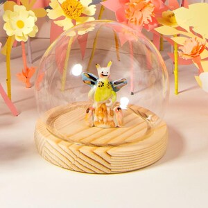 以下が含まれることがあります： ピンク、黄色、白の色合いの紙の花の気まぐれなディスプレイ。木製の台座の上にガラスドームがあり、小さな置物が入っています。このシーンは、春と自然を連想させ、遊び心のある芸術的な美学を持っています。