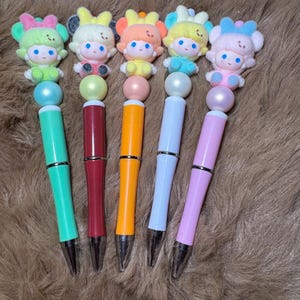 May include: Five colorful pens with decorative doll toppers. Each pen features a different color, including green, red, orange, white, and pink. The doll toppers have various pastel colors and pearl accents. The pens are displayed on a brown, furry surface.