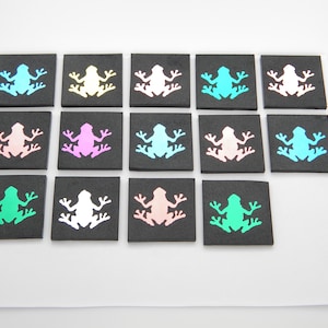 May include: A collection of square, black tiles, each featuring a colorful frog silhouette. The frogs are in various colors, including blue, pink, green, and white. The tiles are arranged on a white surface.