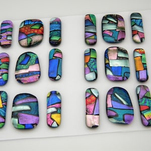 May include: A collection of dichroic glass pendants in assorted shapes, such as rectangles and ovals. The pendants display a mosaic pattern with hues of blue, purple, green, and orange. The glass has a reflective, iridescent finish, creating a shimmering effect.