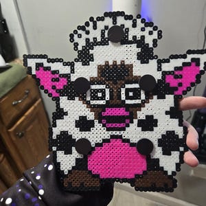 Könnte beinhalten: Ein handgefertigter Kuhmagnet aus bunten Kunststoffperlen. Die Kuh hat einen weißen Körper mit schwarzen Flecken, rosa Ohren und Maul sowie braunen Akzenten. Vier schwarze Magnete sind am Körper der Kuh befestigt.