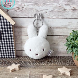 May include: A white plush bunny keychain with a silver-colored bunny-shaped keyring. The bunny has embroidered eyes and a small cross-shaped mouth. The keychain has a small tag that reads "miffy".