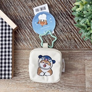 May include: A small, off-white square coin purse with an embroidered cartoon cat wearing a blue and white hat. The purse has a star-shaped clasp and a round tag with a cartoon chef. The background is a wooden surface.