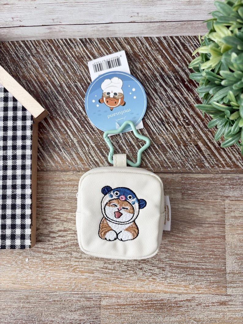 May include: A small, off-white square coin purse with an embroidered cartoon cat wearing a blue and white hat. The purse has a star-shaped clasp and a round tag with a cartoon chef. The background is a wooden surface.