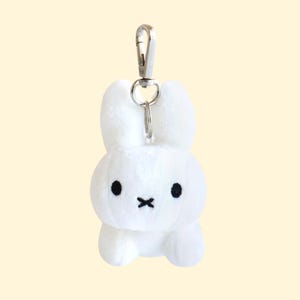 May include: A white plush bunny keychain with black eyes and a stitched mouth. The bunny has a silver metal clip for attaching to keys or bags. The background is a solid light yellow.