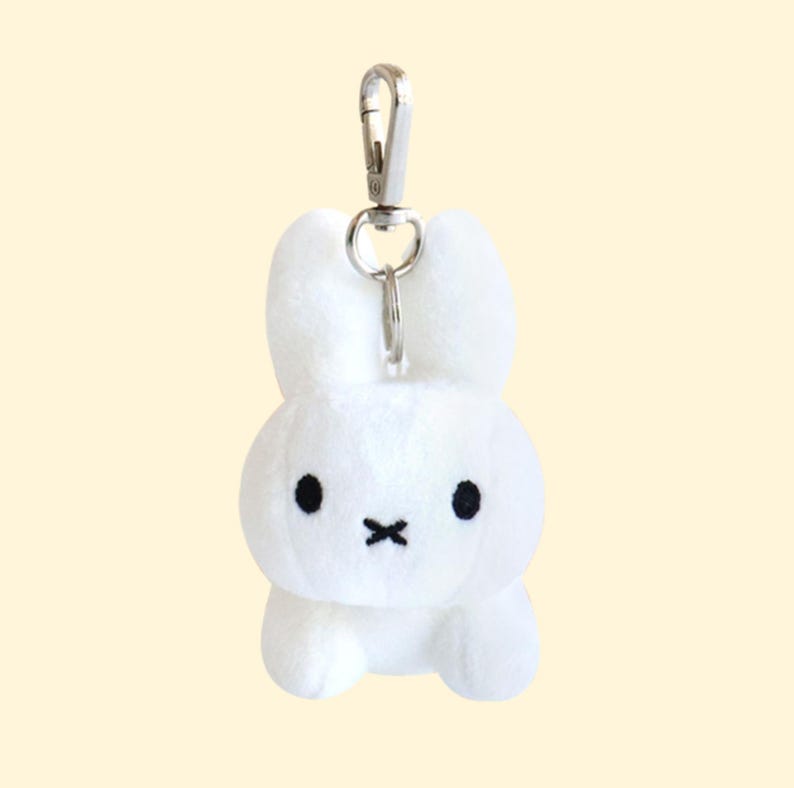 May include: A white plush bunny keychain with black eyes and a stitched mouth. The bunny has a silver metal clip for attaching to keys or bags. The background is a solid light yellow.