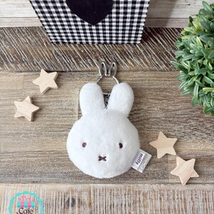 May include: A white plush Miffy keychain with a silver-tone metal keyring. The keychain has embroidered brown eyes and a cross-shaped mouth. A small tag with the name "Miffy" is attached. Wooden star-shaped decorations are scattered around the keychain.