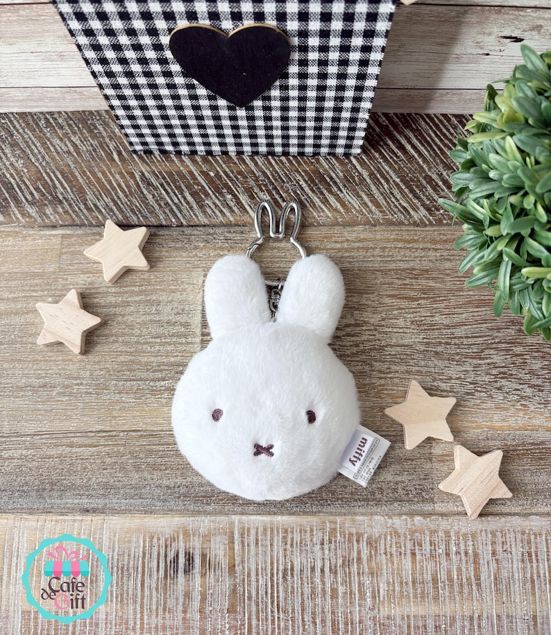 May include: A white plush Miffy keychain with a silver-tone metal keyring. The keychain has embroidered brown eyes and a cross-shaped mouth. A small tag with the name "Miffy" is attached. Wooden star-shaped decorations are scattered around the keychain.