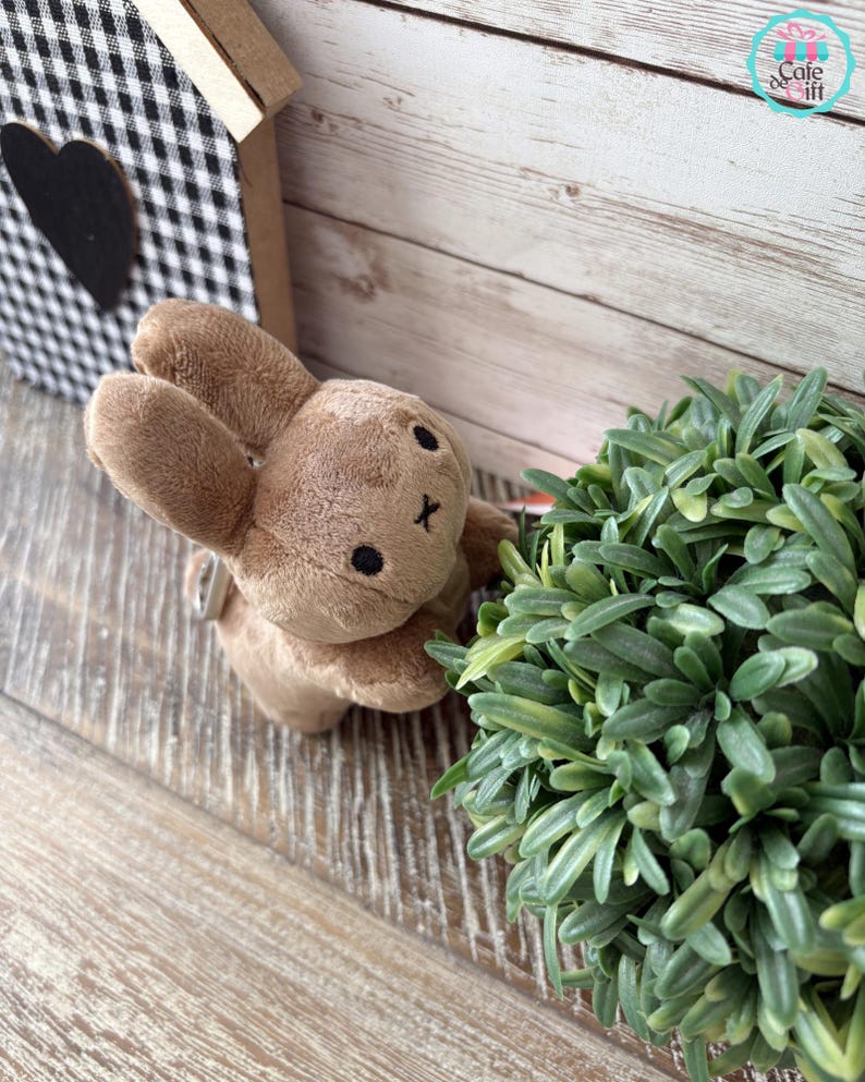 May include: A small, plush brown bunny toy with black eyes and a stitched 'X' for a mouth. The bunny is positioned near a green plant and a decorative wooden house with a black heart. The background is a light-colored wooden surface.
