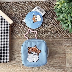 May include: A light blue square pouch with a zipper closure and a pink star-shaped clasp. The pouch features an embroidered image of a white cat wearing a brown hat. A blue circular tag with a cartoon image is attached.