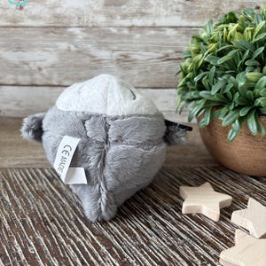 Puede incluir: Un juguete de peluche gris con forma de animal redondo con una cara blanca y ojos negros. El juguete tiene una etiqueta en la parte posterior que dice "CE MADE IN".