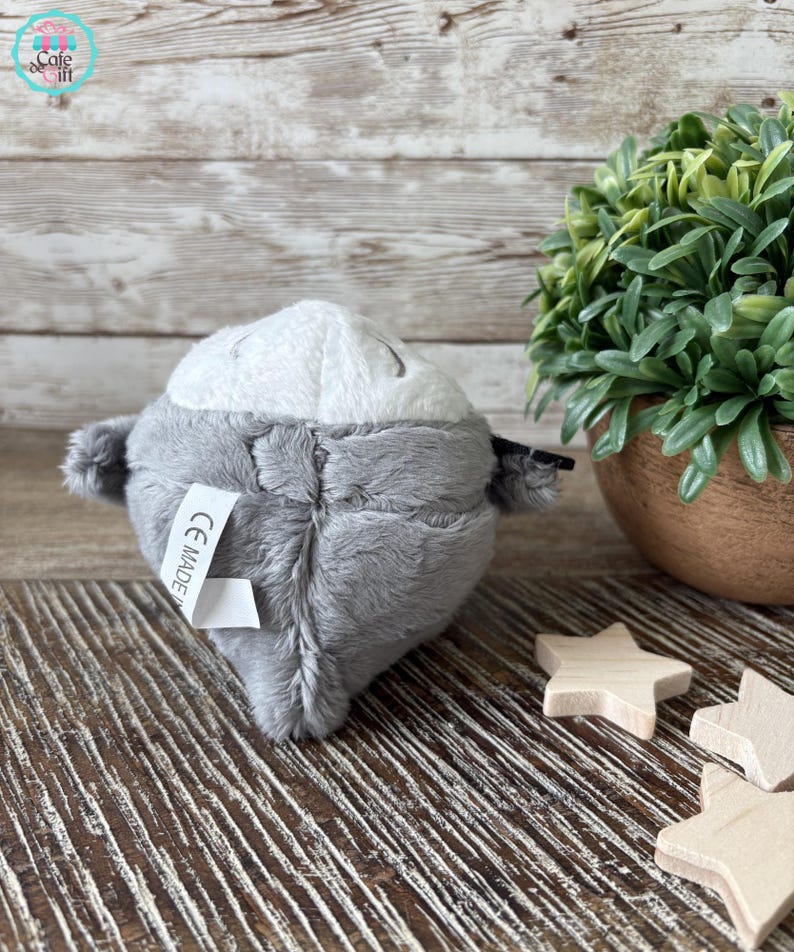 Puede incluir: Un juguete de peluche gris con forma de animal redondo con una cara blanca y ojos negros. El juguete tiene una etiqueta en la parte posterior que dice "CE MADE IN".