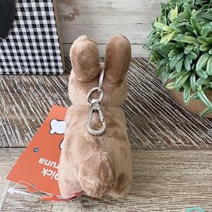 May include: A plush, light brown bunny keychain with a silver clasp. The bunny has long ears and a fluffy tail. An orange tag with the text "pick tuna" is attached. The background includes a black and white checkered pattern and a potted plant.