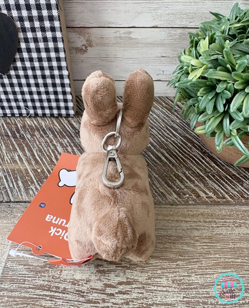 May include: A plush, light brown bunny keychain with a silver clasp. The bunny has long ears and a fluffy tail. An orange tag with the text "pick tuna" is attached. The background includes a black and white checkered pattern and a potted plant.