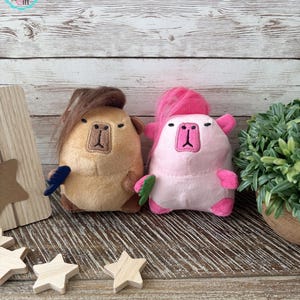 May include: Two plush toys shaped like capybaras. One is tan with brown hair and a blue feather, the other is pink with pink hair and a green leaf. The toys have embroidered faces and are on a wooden surface.