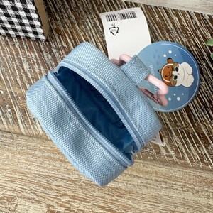 May include: A light blue zippered pouch with a matching interior. A round blue tag with a cartoon dog illustration is visible. The pouch is open, revealing the interior. A pink strap is attached to the pouch.