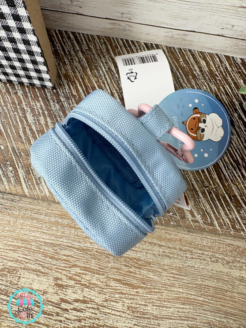 May include: A light blue zippered pouch with a matching interior. A round blue tag with a cartoon dog illustration is visible. The pouch is open, revealing the interior. A pink strap is attached to the pouch.