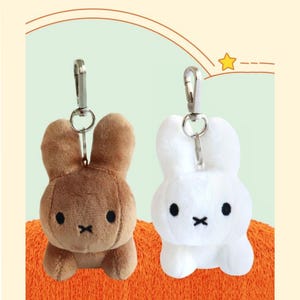 May include: Two plush bunny keychains, one brown and one white, each with a silver clip. The bunnies have black eyes and a black 'X' mouth. They are set against an orange and light green background.