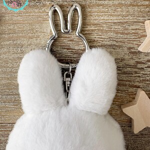 May include: A white fluffy bunny keychain with a silver-colored metal bunny ear design. The keychain has a silver clasp and is made of soft, plush material. The background is a light brown wooden surface.