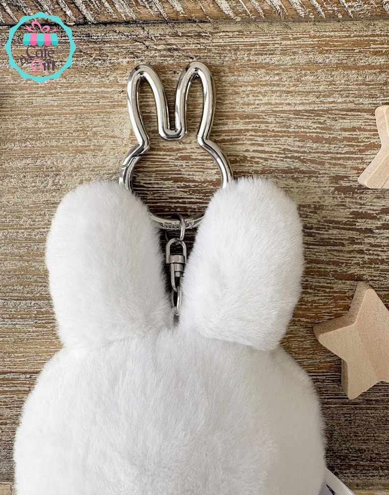 May include: A white fluffy bunny keychain with a silver-colored metal bunny ear design. The keychain has a silver clasp and is made of soft, plush material. The background is a light brown wooden surface.