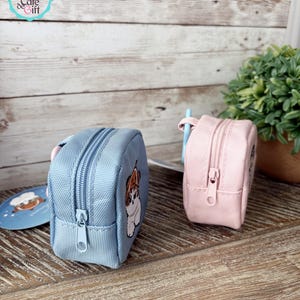 May include: Two small, square zippered pouches. One is light blue with a dog graphic, the other is pink. Both have a matching zipper and a small loop. The pouches are on a wooden surface.