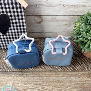 May include: Two small, square, blue pouches with star-shaped clasps. One clasp is white, the other is pink. The pouches have the word "mofusand" printed in a light color. The background includes a wooden surface and a small decorative house.