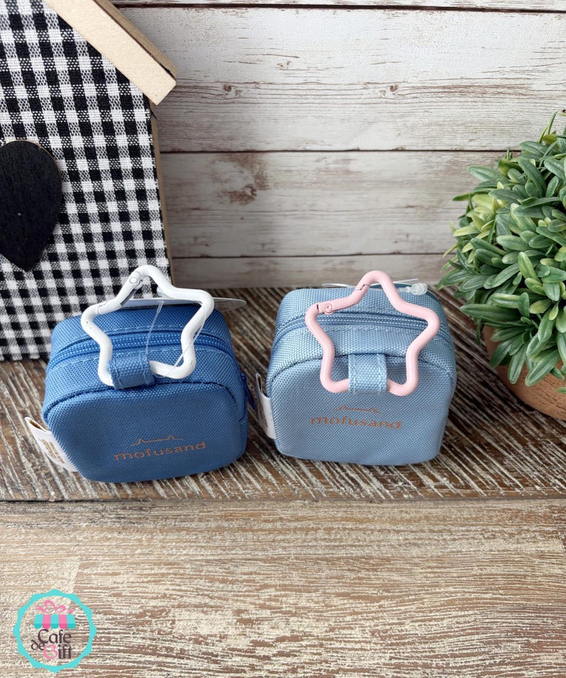 May include: Two small, square, blue pouches with star-shaped clasps. One clasp is white, the other is pink. The pouches have the word "mofusand" printed in a light color. The background includes a wooden surface and a small decorative house.