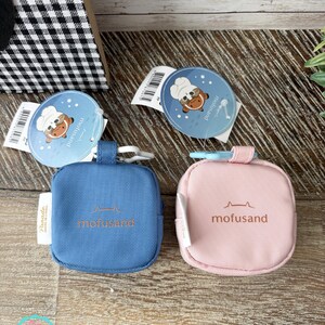 May include: Two small, square pouches, one blue and one pink, with the word "mofusand" printed on them. Each pouch has a small loop and a tag with a cartoon cat wearing a chef's hat. The pouches are on a wooden surface.