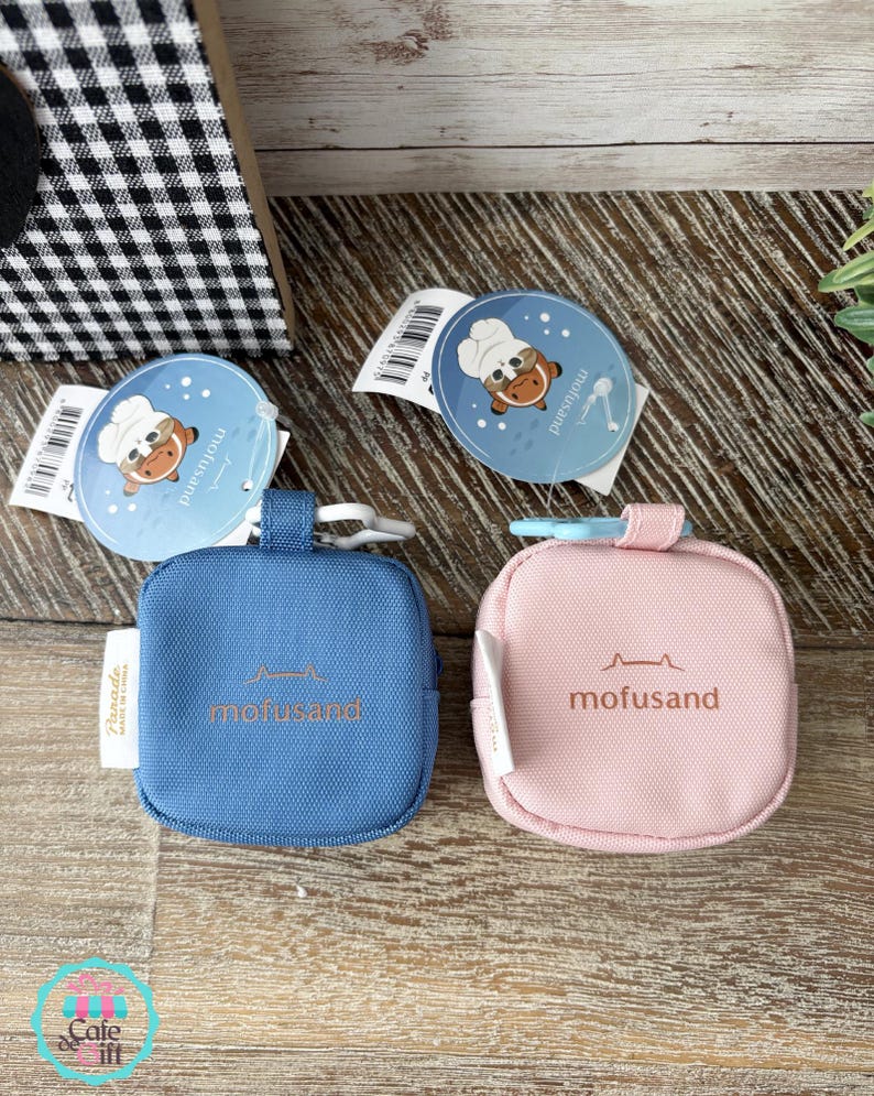 May include: Two small, square pouches, one blue and one pink, with the word "mofusand" printed on them. Each pouch has a small loop and a tag with a cartoon cat wearing a chef's hat. The pouches are on a wooden surface.