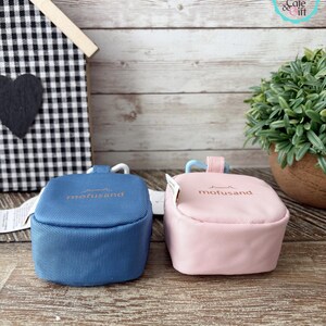 May include: Two small, square pouches in blue and pink, side-by-side. Each has a curved handle and the word "mofusand" printed in gold. The pouches are on a wooden surface, with a small house-shaped decoration in the background.