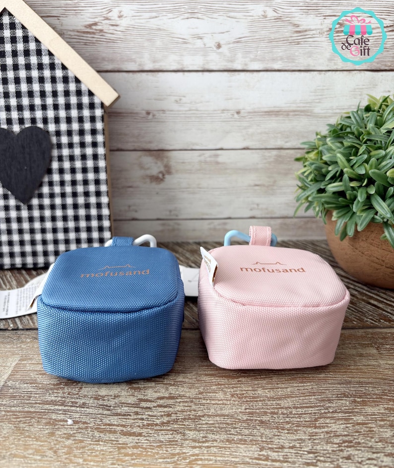 May include: Two small, square pouches in blue and pink, side-by-side. Each has a curved handle and the word "mofusand" printed in gold. The pouches are on a wooden surface, with a small house-shaped decoration in the background.